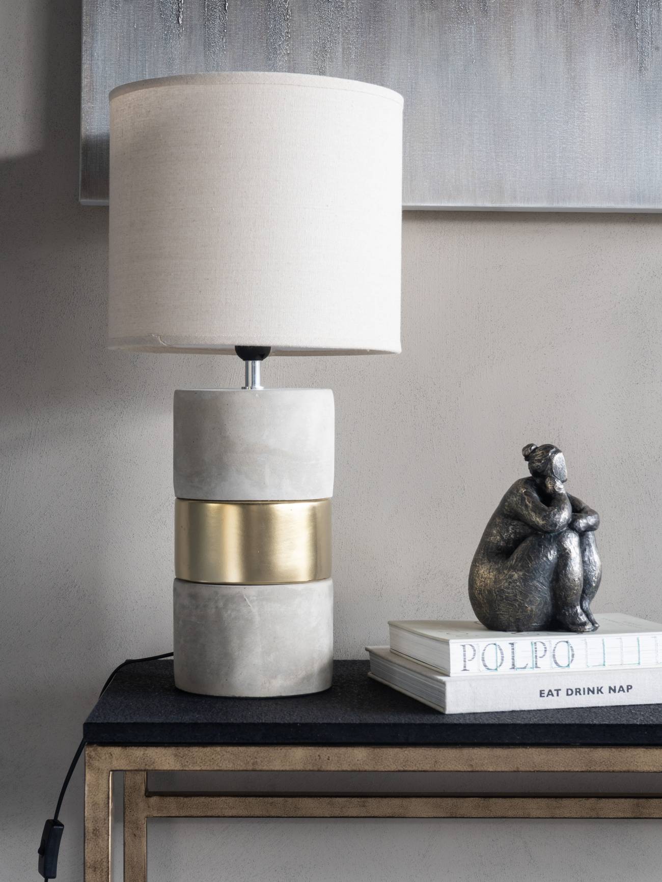 Table lamp with a concrete base and gold band on a dark surface with books and a sculpture.